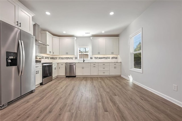 kitchen featuring appliances with stainless steel finishes, backsplash, wall chimney exhaust hood, light hardwood / wood-style flooring, and white cabinetry