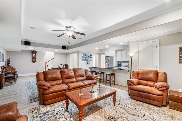 living room featuring ceiling fan, baseboards, stairs, light wood-style flooring, and a raised ceiling