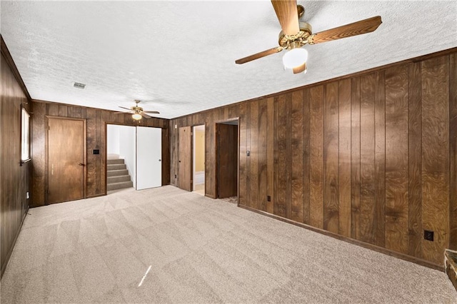 carpeted empty room with visible vents, a ceiling fan, a textured ceiling, stairway, and wood walls