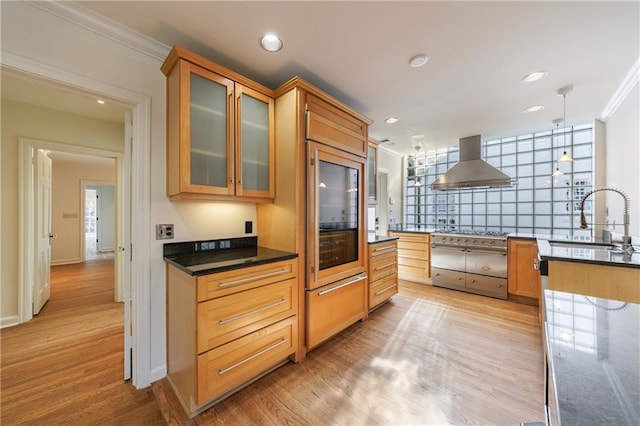 kitchen with wall chimney range hood, crown molding, sink, hanging light fixtures, and light hardwood / wood-style flooring