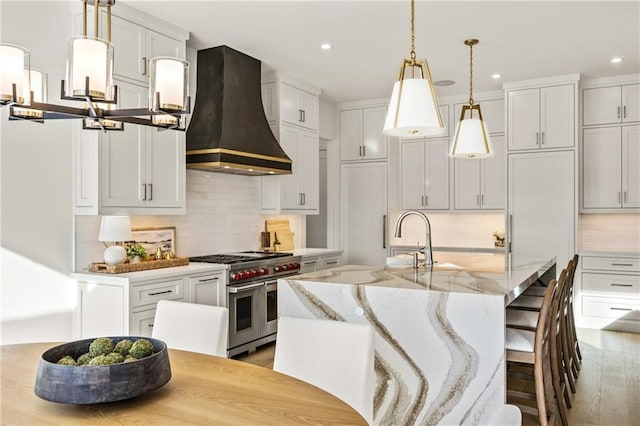 kitchen with light stone counters, custom exhaust hood, double oven range, an island with sink, and decorative light fixtures