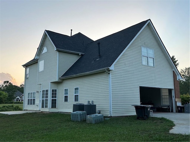 back house at dusk with a garage, cooling unit, and a lawn