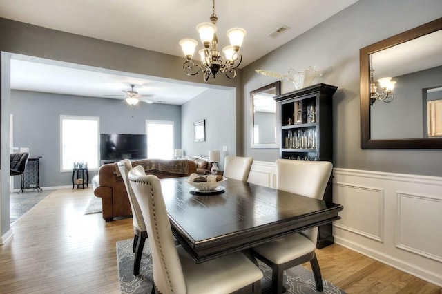 dining room with visible vents, wainscoting, light wood-style floors, a decorative wall, and ceiling fan with notable chandelier