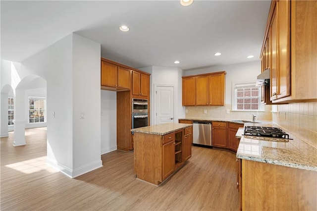 kitchen with brown cabinets, light wood-style flooring, arched walkways, appliances with stainless steel finishes, and decorative backsplash