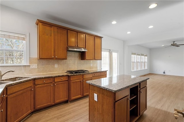 kitchen with under cabinet range hood, decorative backsplash, brown cabinets, light wood-style floors, and a sink