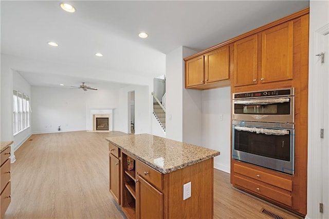 kitchen with a fireplace with flush hearth, brown cabinets, light wood-style floors, stainless steel double oven, and a ceiling fan