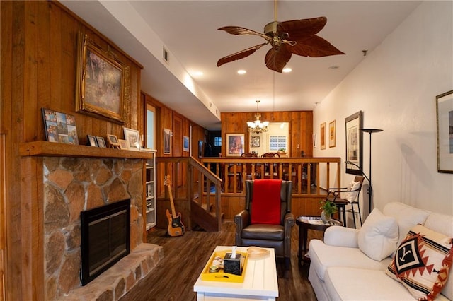 living room with a stone fireplace, dark hardwood / wood-style floors, ceiling fan with notable chandelier, and wooden walls