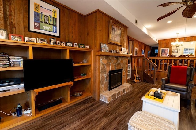 living room featuring a stone fireplace, built in shelves, dark hardwood / wood-style flooring, ceiling fan with notable chandelier, and wood walls