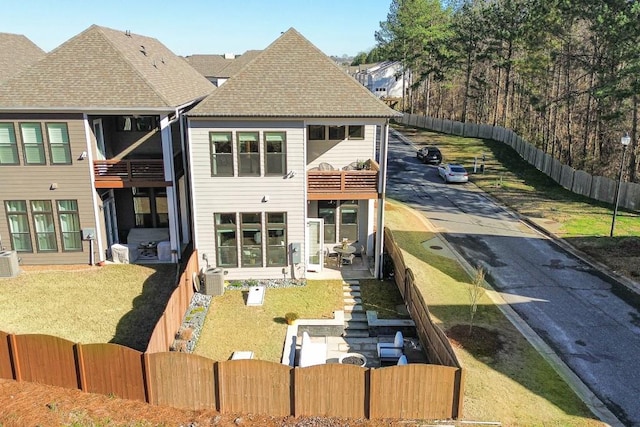 back of house with a balcony, a fenced front yard, and roof with shingles