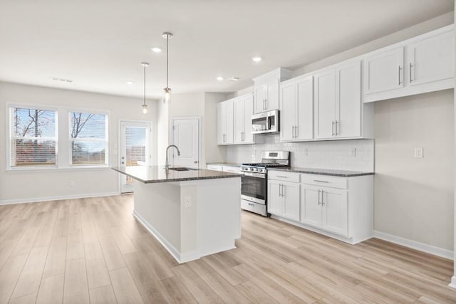 kitchen featuring white cabinetry, hanging light fixtures, an island with sink, stainless steel appliances, and dark stone counters