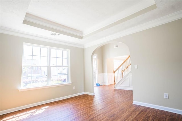empty room featuring wood finished floors, arched walkways, a raised ceiling, ornamental molding, and visible vents