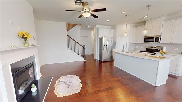 kitchen featuring appliances with stainless steel finishes, a sink, a glass covered fireplace, an island with sink, and a ceiling fan