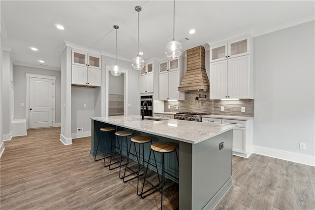 kitchen with custom exhaust hood, white cabinets, crown molding, sink, and an island with sink