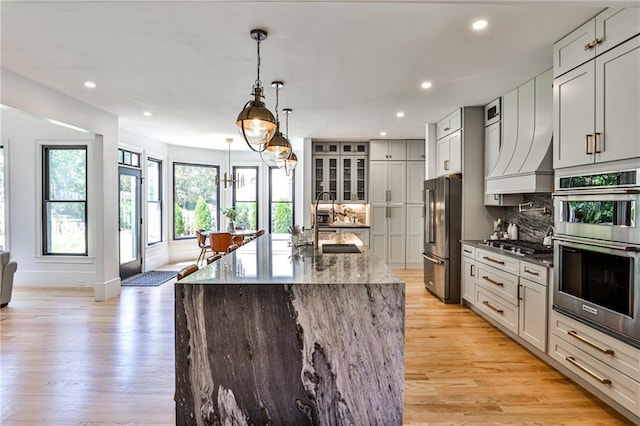 kitchen with appliances with stainless steel finishes, hanging light fixtures, an island with sink, light stone countertops, and tasteful backsplash