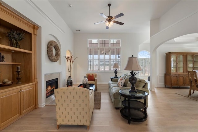 living area featuring light wood-type flooring, arched walkways, a warm lit fireplace, and ceiling fan