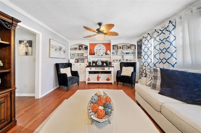 living area with built in shelves, wood finished floors, baseboards, ceiling fan, and crown molding