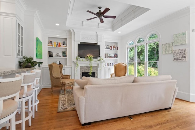 living room with a high end fireplace, a tray ceiling, light hardwood / wood-style flooring, ornamental molding, and ceiling fan