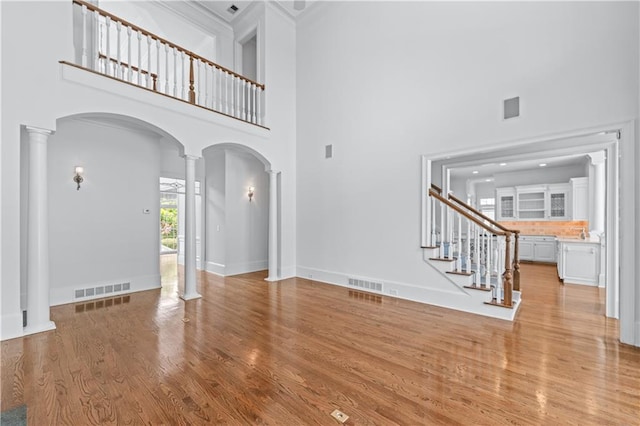 unfurnished living room featuring decorative columns, visible vents, arched walkways, light wood-type flooring, and a high ceiling