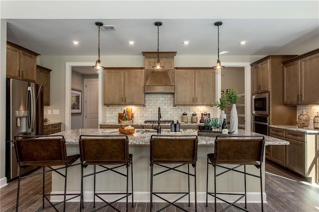 kitchen featuring light stone countertops, custom range hood, stainless steel appliances, pendant lighting, and a center island with sink