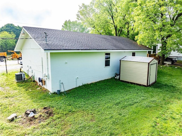 rear view of property with a shingled roof, a storage shed, central AC unit, a yard, and an outbuilding
