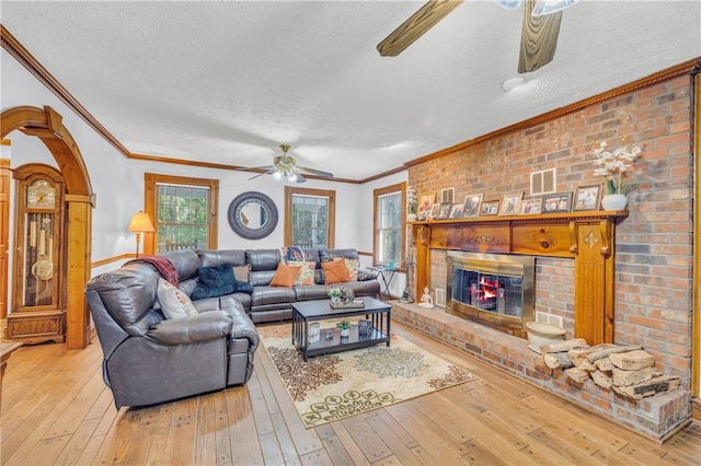 living room featuring a textured ceiling, light hardwood / wood-style flooring, ornamental molding, a brick fireplace, and ceiling fan