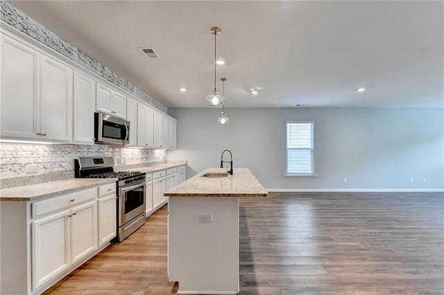 kitchen with light stone counters, a kitchen island with sink, stainless steel appliances, white cabinetry, and decorative light fixtures