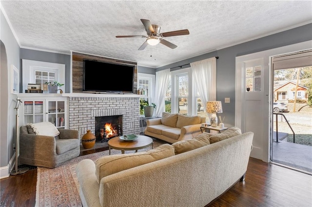 living room with a brick fireplace, ornamental molding, a textured ceiling, ceiling fan, and dark wood-type flooring