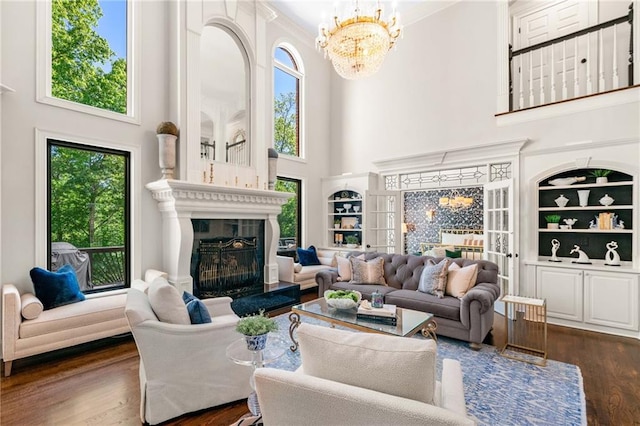 living area with a high ceiling, dark wood-type flooring, a fireplace, a chandelier, and built in shelves
