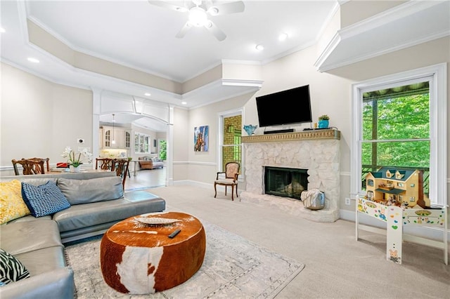 living room featuring a ceiling fan, carpet flooring, crown molding, a stone fireplace, and baseboards