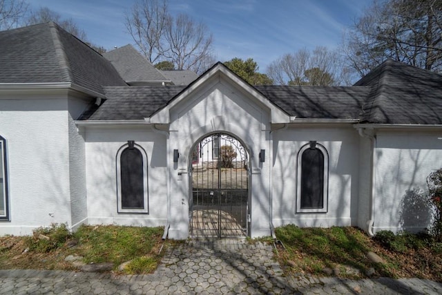 view of exterior entry featuring a gate, stucco siding, and a shingled roof