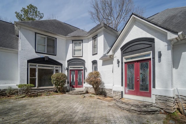 entrance to property with french doors, stone siding, roof with shingles, and stucco siding