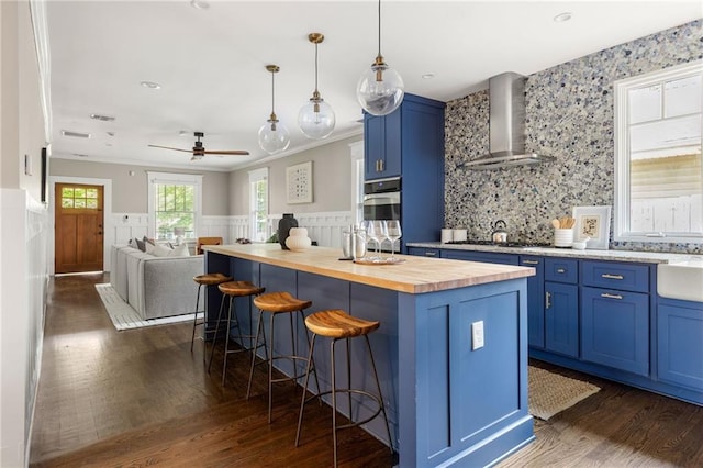 kitchen with butcher block countertops, blue cabinets, a kitchen bar, wall chimney range hood, and dark wood-style floors