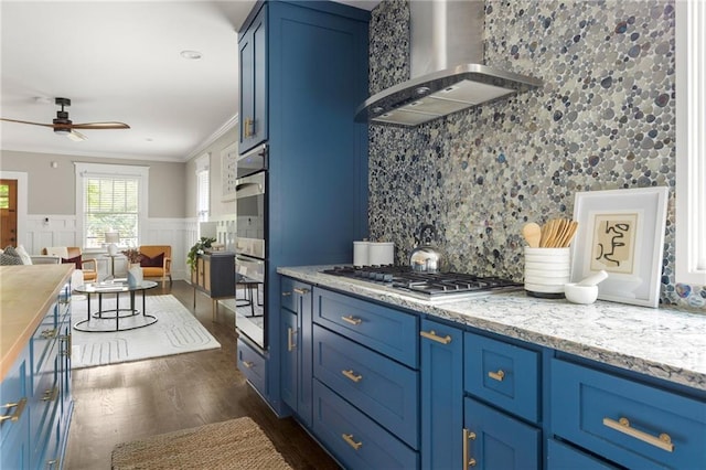 kitchen with wall chimney range hood, stainless steel appliances, blue cabinets, ornamental molding, and dark wood-type flooring