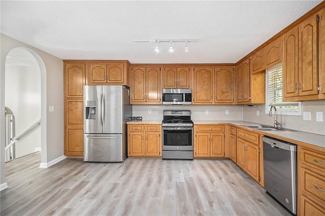 kitchen featuring appliances with stainless steel finishes, arched walkways, brown cabinets, a sink, and light wood-style floors
