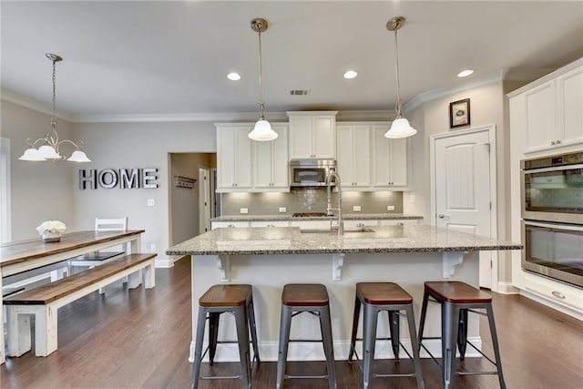 kitchen featuring an island with sink, stainless steel appliances, decorative light fixtures, white cabinetry, and light stone counters