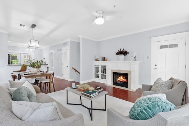 living area featuring a ceiling fan, a fireplace with flush hearth, wood finished floors, visible vents, and ornamental molding