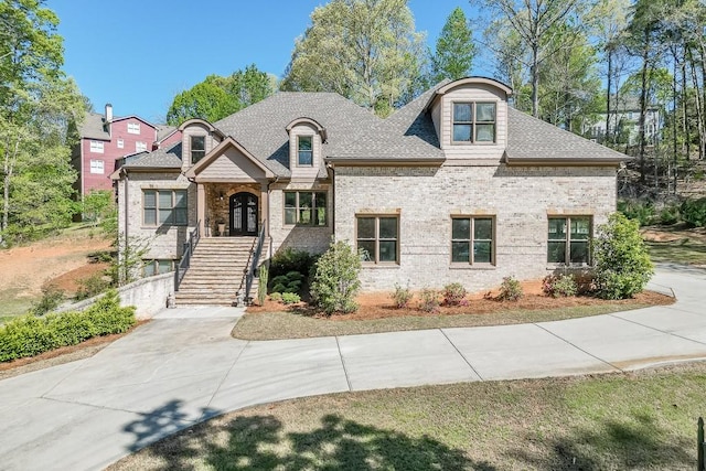 french country inspired facade with brick siding, a shingled roof, and stairs