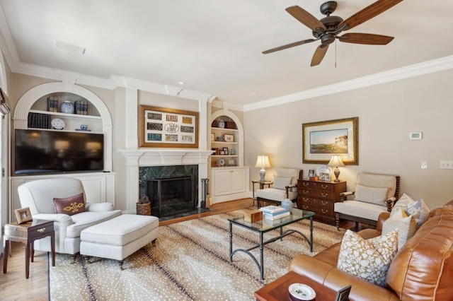 living room with ornamental molding, built in shelves, light wood-type flooring, a high end fireplace, and ceiling fan