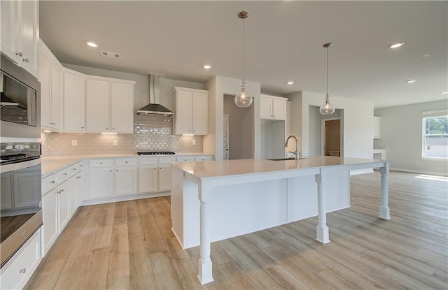 kitchen featuring sink, white cabinetry, decorative light fixtures, an island with sink, and wall chimney range hood