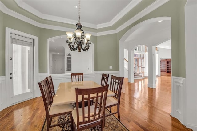 dining area featuring a wainscoted wall, stairway, light wood finished floors, a raised ceiling, and an inviting chandelier