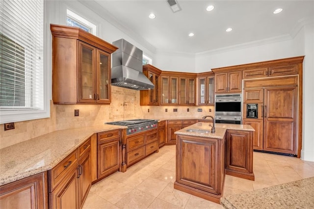 kitchen featuring light stone counters, a sink, a center island with sink, wall chimney exhaust hood, and glass insert cabinets