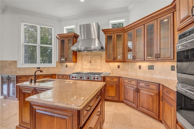 kitchen featuring light stone counters, a sink, brown cabinets, wall chimney exhaust hood, and glass insert cabinets