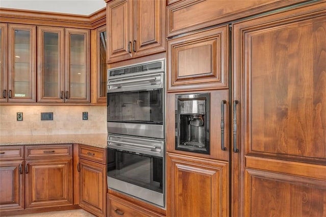 kitchen with decorative backsplash, glass insert cabinets, light stone counters, brown cabinets, and double oven