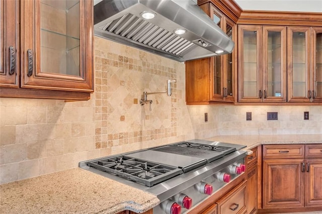 kitchen featuring glass insert cabinets, brown cabinets, light stone counters, ventilation hood, and stainless steel gas cooktop