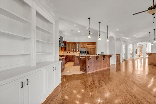 kitchen featuring brown cabinets, a spacious island, light countertops, wall chimney range hood, and a kitchen bar