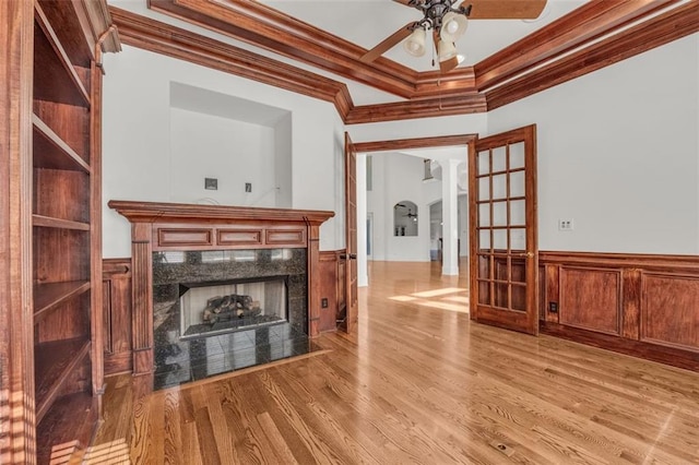 unfurnished living room featuring a ceiling fan, wainscoting, crown molding, light wood-type flooring, and a fireplace