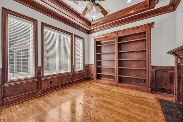 interior space featuring light wood-style flooring, a wainscoted wall, a ceiling fan, visible vents, and crown molding