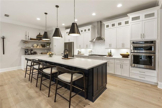 kitchen with a kitchen island with sink, hanging light fixtures, white cabinetry, stainless steel appliances, and wall chimney exhaust hood