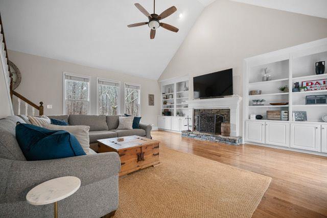 living room with high vaulted ceiling, a stone fireplace, built in shelves, a ceiling fan, and light wood-type flooring