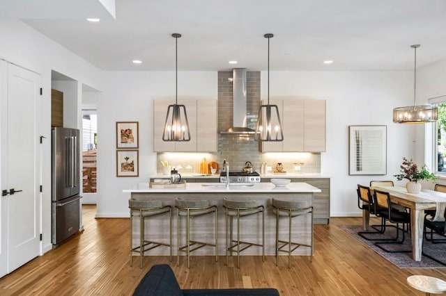 kitchen with decorative light fixtures, sink, a kitchen island with sink, high end fridge, and wall chimney exhaust hood